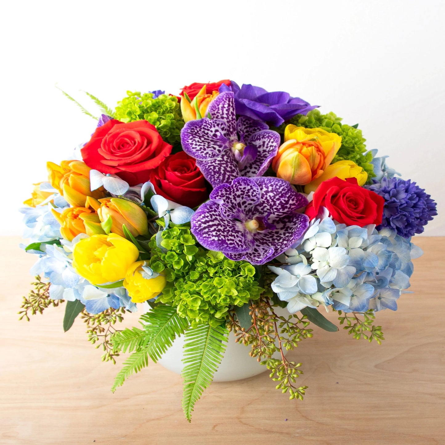This brightly colored arrangement features red roses, orange and yellow tulips, blue hydrangea, and purple vanda orchids.  Note: exact flowers will vary. Our team will always choose the freshest, most unique seasonal flower.