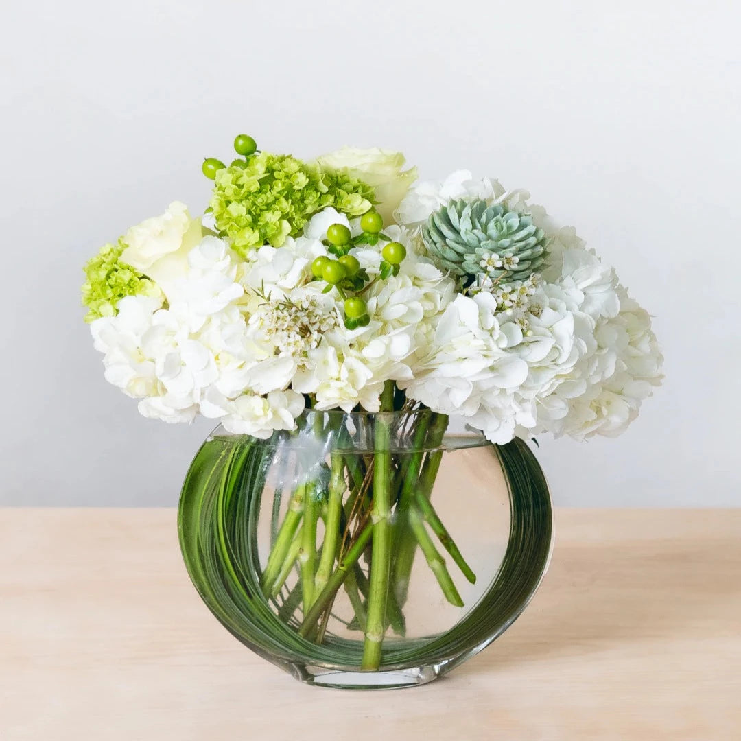 A design of whites and greens that never goes out of style. Beautiful hydrangeas and Elegant white roses are accented with hypericum berries and an echeveria in a flat fish bowl.