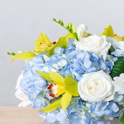 This design is classic and timeless, just like California. In a white glass vase, light blue hydrangea, green cymbidium orchids, and white roses are enhanced with white freesia.