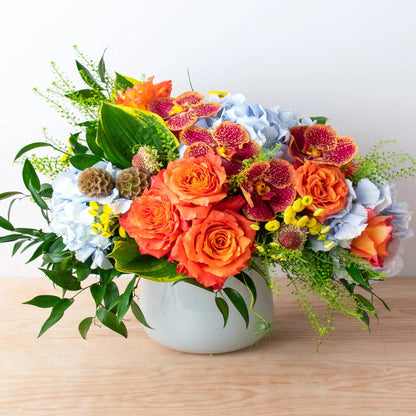 This one-of-a-kind floral pattern transports you to a summer sunset.  orchids, Verea blue hydrangea, and garden roses are among the novelties. In a huge white glass vase, yellow Matricaria, scabiosa pods, green bell Thlaspi, and hosta leaves are accented.