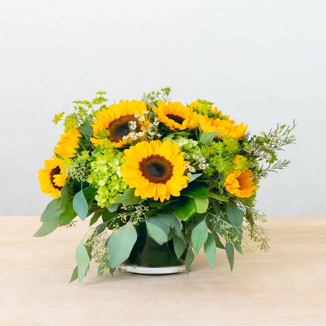 Local sunflowers are complemented with eucalyptus and other textural elements. This arrangement, which is designed in a glass, leaf-lined cylinder, is ideal for brightening up any environment.