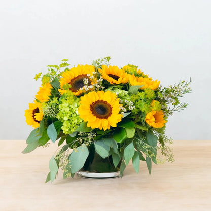 Local sunflowers are complemented with eucalyptus and other textural elements. This arrangement, which is designed in a glass, leaf-lined cylinder, is ideal for brightening up any environment.